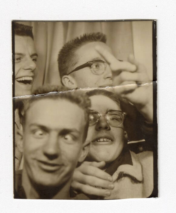 Black and white photograph of four men in a photobooth. Man in the foreground is making a funny face.
